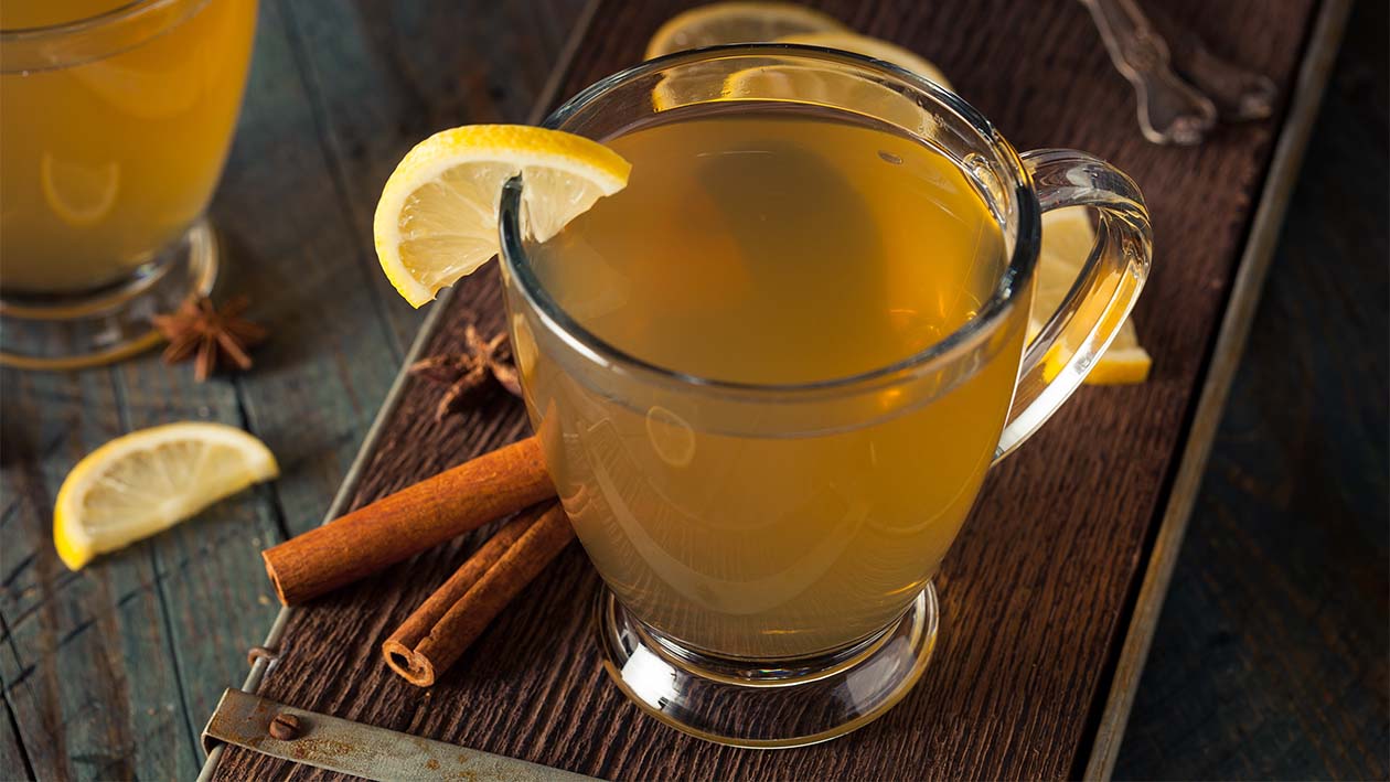 Gelbes Getränk in durchsichtiger Tasse mit Zitronenscheibe und Zimtstangen