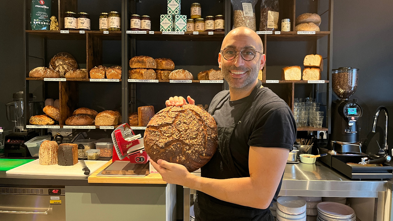 „Lucky“ Theiss hält ein Brot hinter der Theke in der Hand