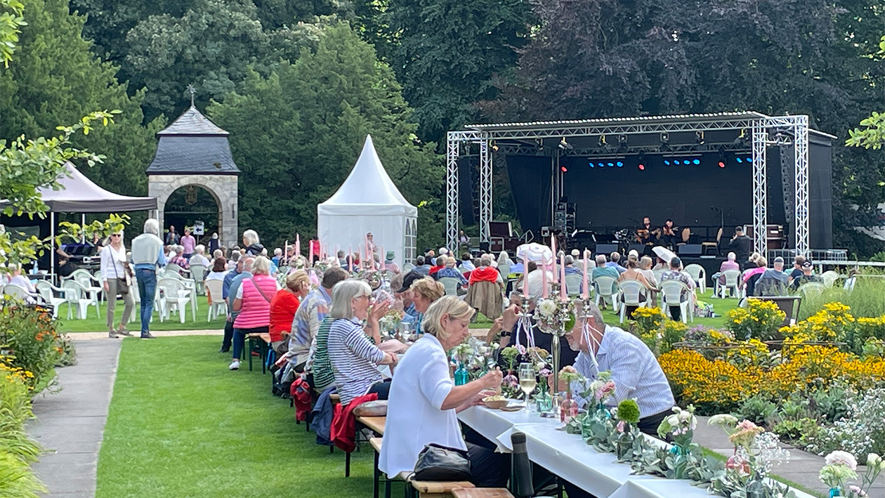 Viele Menschen sitzen an einer Langen Tafel, im Hintergrund ist eine Bühne zu sehen