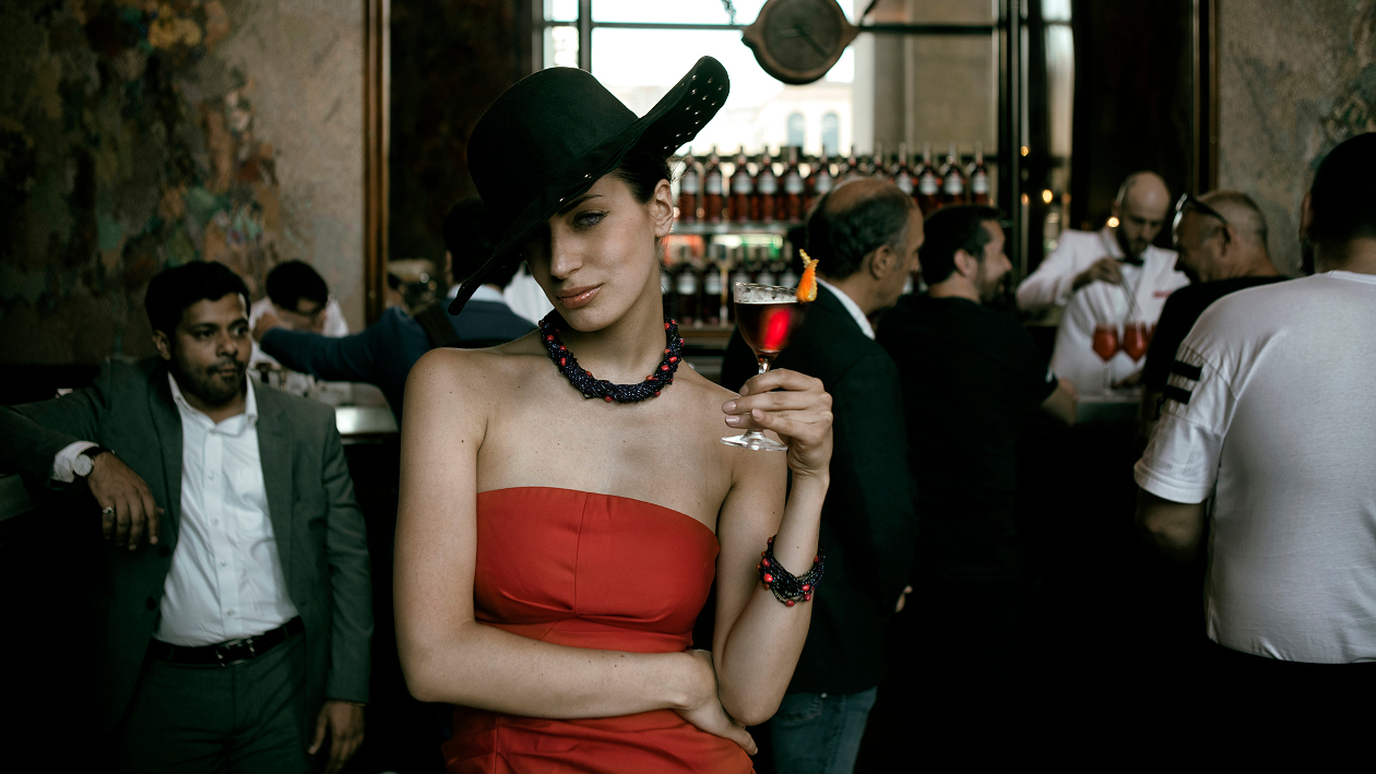 Eine Frau, gekleidet in einem roten Kleid und einem schwarzen Hut, steht in einer Bar und hält ein Glas Rotwein fest