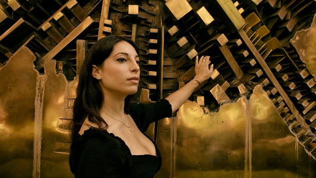 Eine Person, gekleidet in einem schwarzen Kleid, steht vor einem goldenen metallischen Hintergrund
