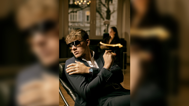 Ein Mann in einem Anzug sitzt auf einem Lederstuhl, er trägt Sonnenbrille und hält eine Zigarre fest