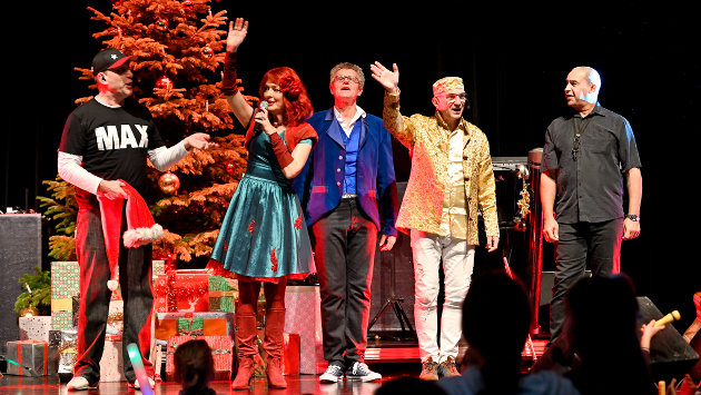 Mehrere Menschen stehen auf einer Bühne, im Hintergrund steht ein Weihnachtsbaum mit Geschenken drunter