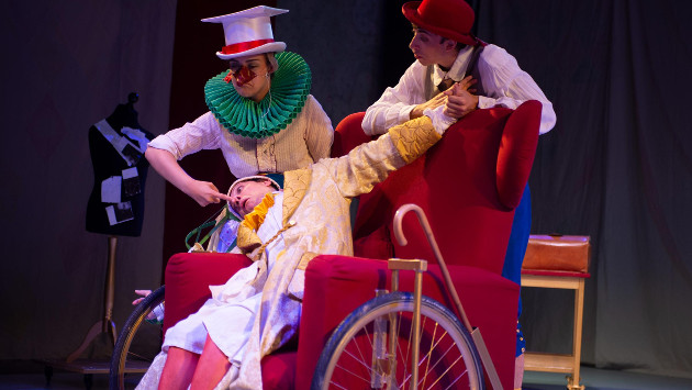 Eine Szene aus dem Theaterstück "Der eingebildete Kranke", zwei Schauspieler stehen hinter einem Schauspieler im Rollstuhl auf der Bühne