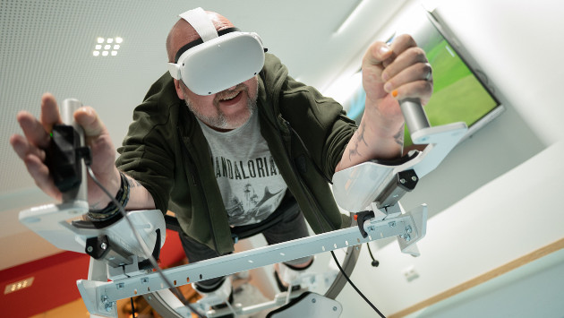 Eine Person liegt auf einen VR-System und spielt mit einer VR-Brille ein Spiel