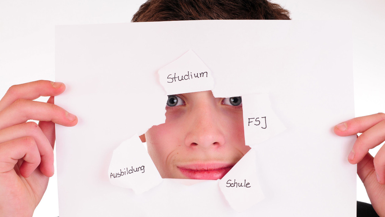 Ein junge hält ein blatt Papier vor sein Gesicht und schaut durch ein herausgerissenes Loch. Auf dem Papier stehen vier Wörter, Studium, FSJ, Schule und Ausbildung.