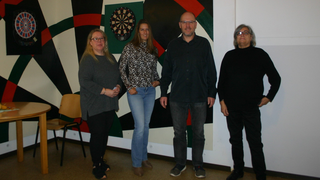 Iris Gormanns, Jeannine Ehlen, Christoph Föhles und Sigrid Arroyo stehen im Café Emmaus vor einer Wand, welche mit einer großen Dartscheibe bemalt ist.